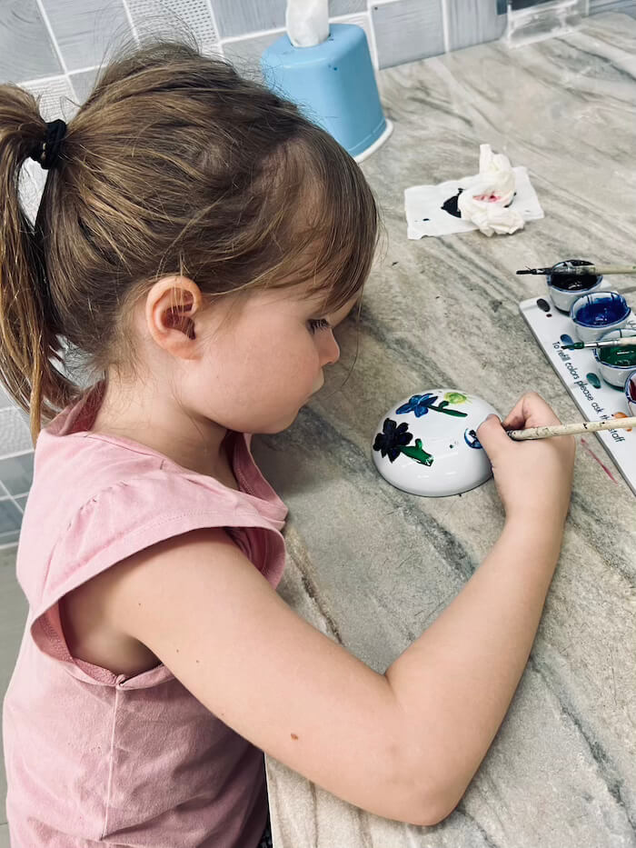 Painting Ceramic Bowls at Dhanabadee Museum A young girl with a ponytail wearing a pink shirt carefully paints blue floral designs onto a small white ceramic bowl at a workshop table.