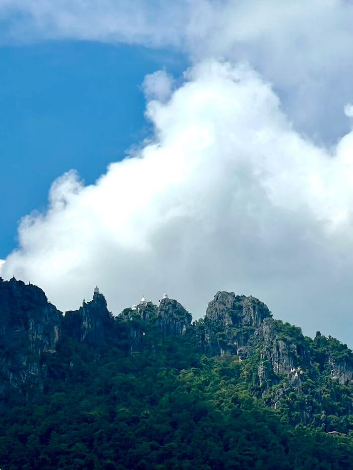 A distant view of a jagged, forested mountain ridge with tiny white pagodas visible on the highest peaks under a large, dramatic white cloud.