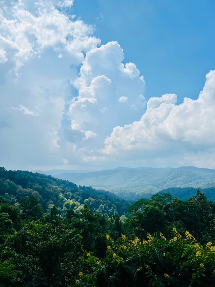 A wide panoramic view of lush green mountain ridges under a bright blue sky with dramatic white clouds at Doi Khun Tan National Park.