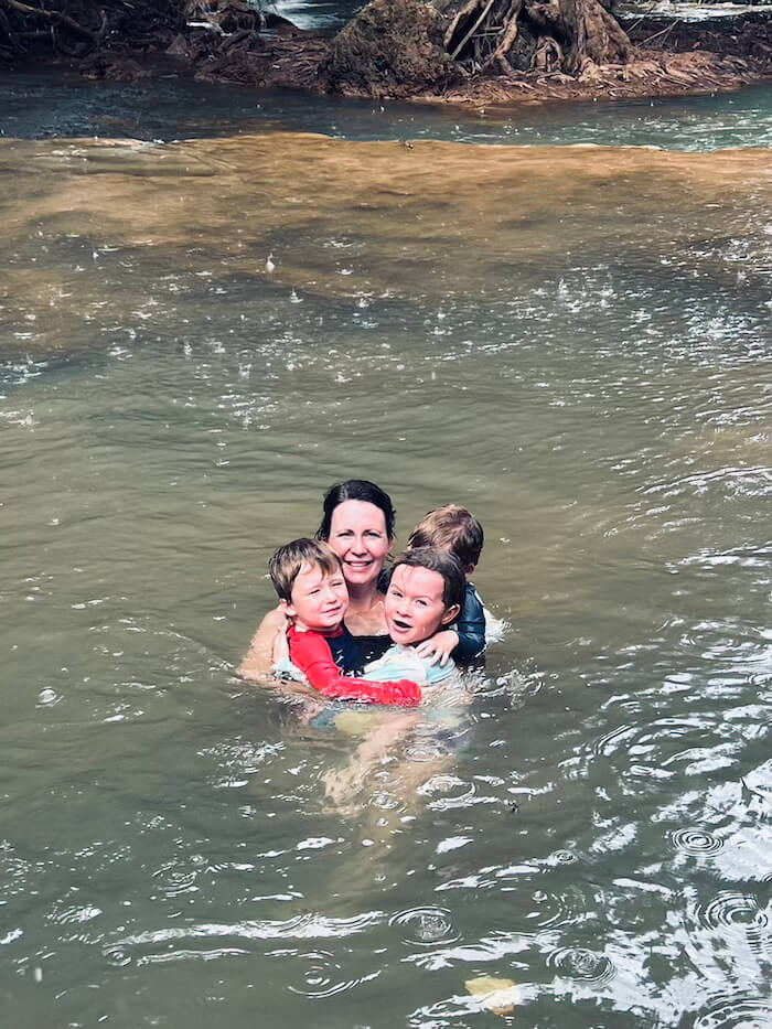 A woman and three young children hugging and smiling while waist-deep in a jungle pool at Thansawan Waterfall during a light rain shower.