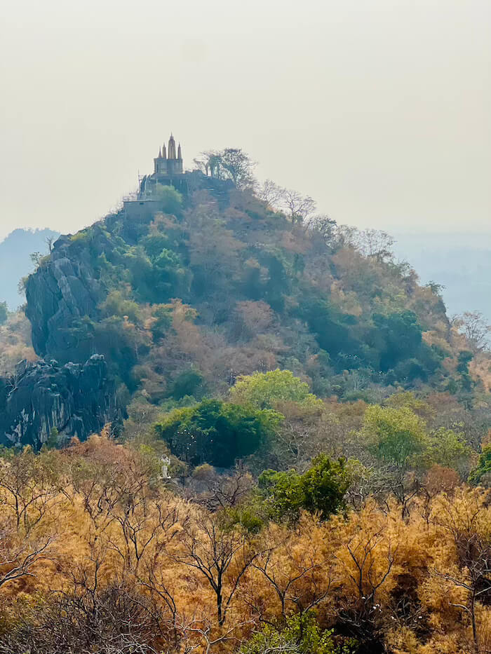 A distant view of a temple structure perched atop a sheer limestone ridge surrounded by dry forest and haze in Ratchaburi.
