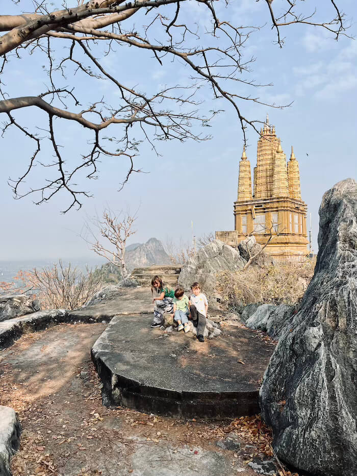Three children sit on a rocky limestone path leading toward a golden Thai pagoda at the Huppha Sawan religious site.