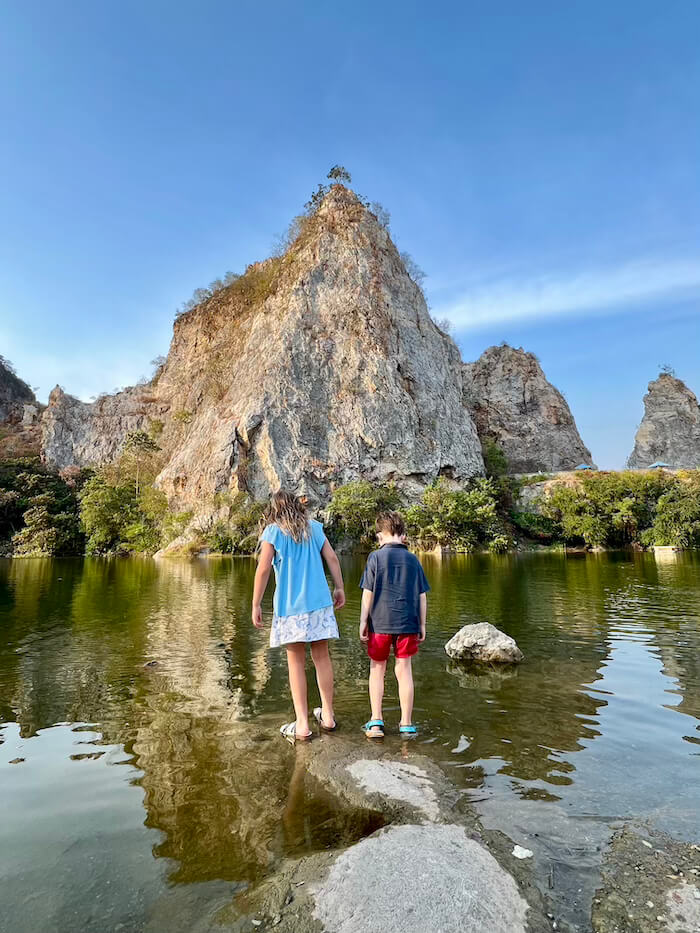 Two children standing on stones at the edge of a green lake in Khao Ngu Stone Park, with a massive limestone mountain in the background.