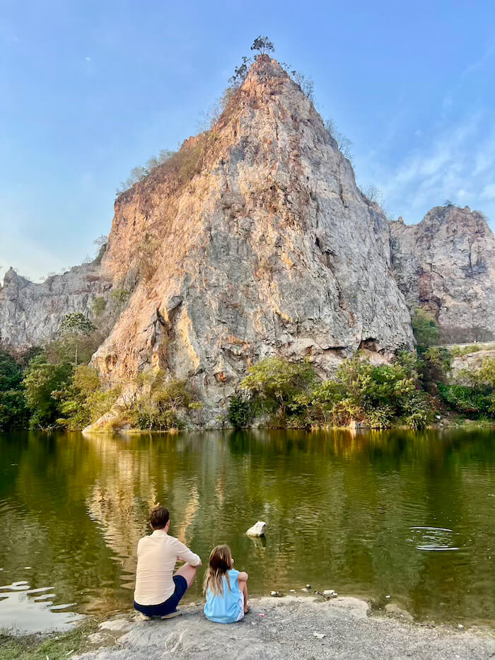 A father and daughter sit by the edge of a green lake at Khao Ngu Stone Park in Ratchaburi, looking up at a towering limestone cliff under a blue sky.