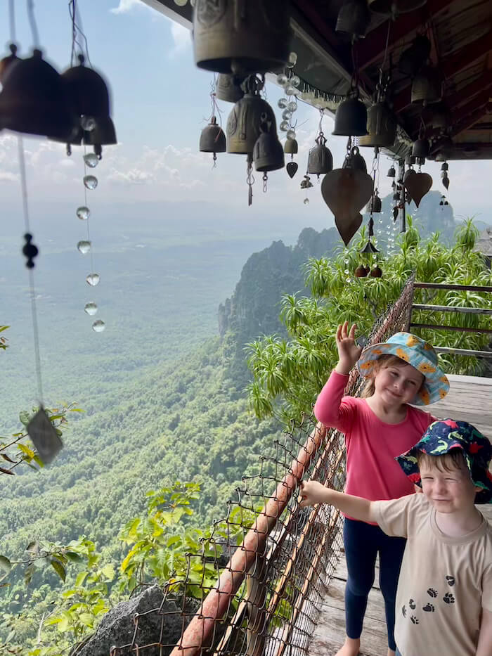 Two young children standing on a wooden temple balcony decorated with small bronze bells, overlooking a deep forested valley.