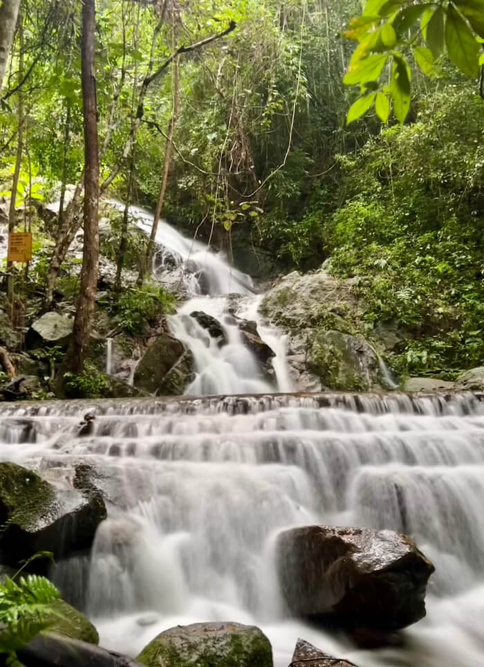 A multi-tiered waterfall cascading over mossy rocks in a dense, green jungle setting with vines hanging from the canopy.