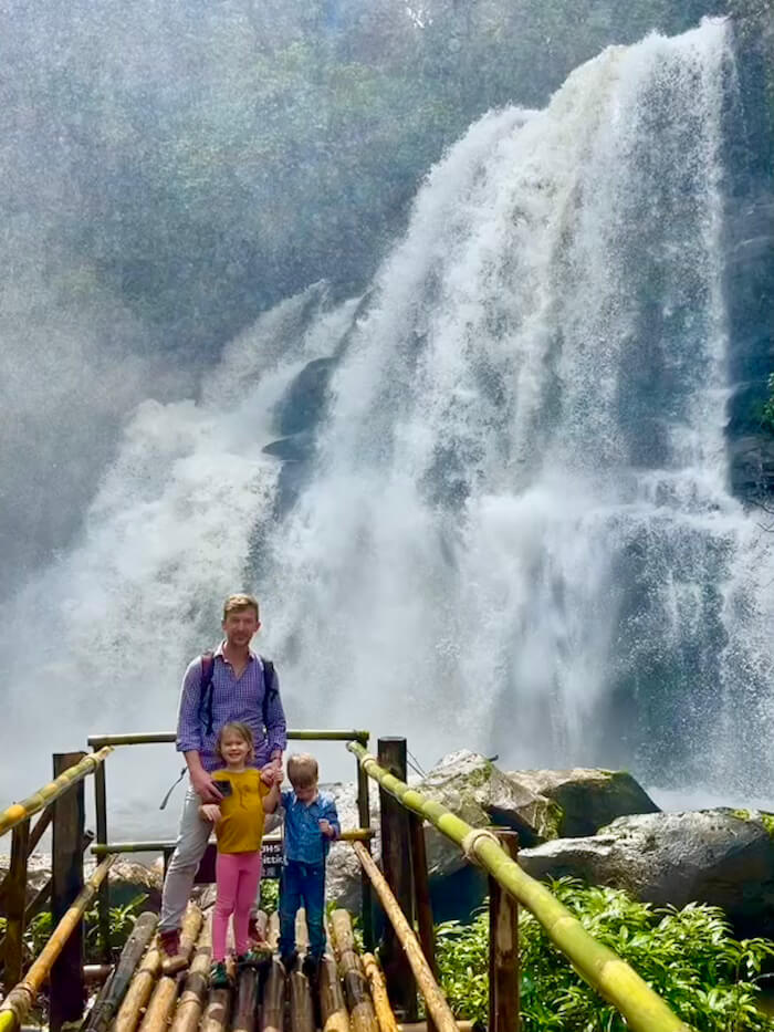 A man and two young children standing on a rustic bamboo bridge in front of a massive, misty waterfall on the Pha Dok Siew Nature Trail, Doi Inthanon.