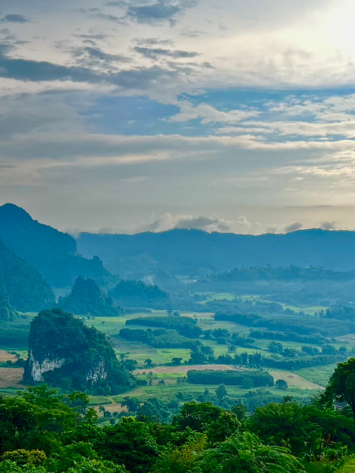 A high-angle view of the Phu Langka valley at sunrise, with the Pha Chang Noi limestone karst in the foreground and layers of blue mountains in the misty distance.