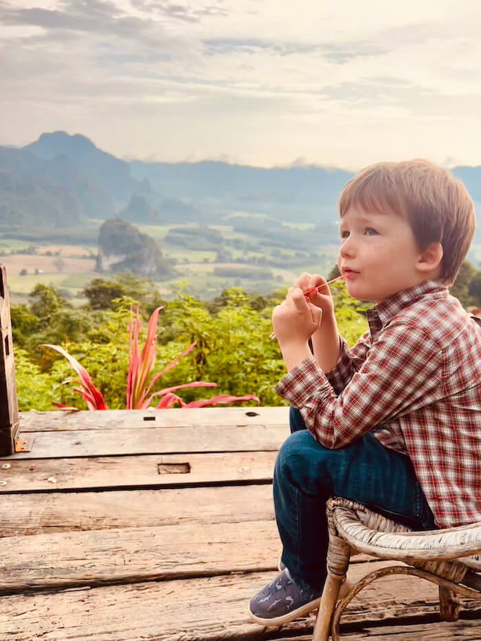 A young boy in a plaid shirt sitting on a wooden deck, looking out over the limestone karsts and misty valley of Phu Langka in Northern Thailand.