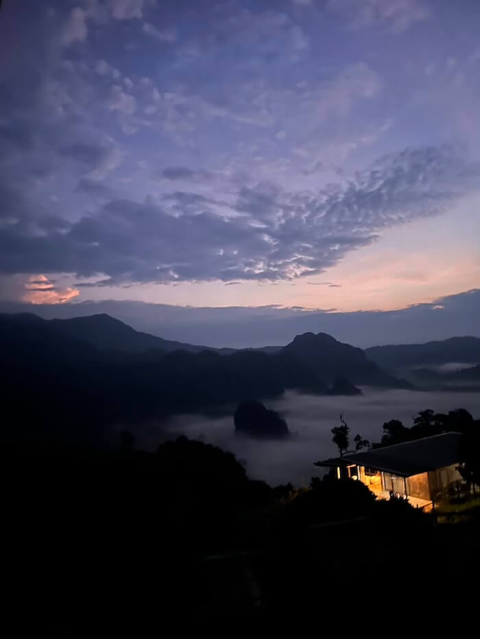 Pre-Dawn Mist at Phu Langka A pre-dawn view from a Phu Langka ridge homestay showing a lit cabin overlooking a valley filled with thick white mist under a dark blue twilight sky.
