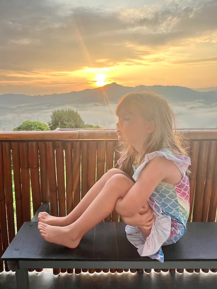 Sunrise Reflection at Phu Langka A young girl sitting quietly on a bamboo balcony, watching the sun rise over a mist-filled valley in Phu Langka, Thailand.