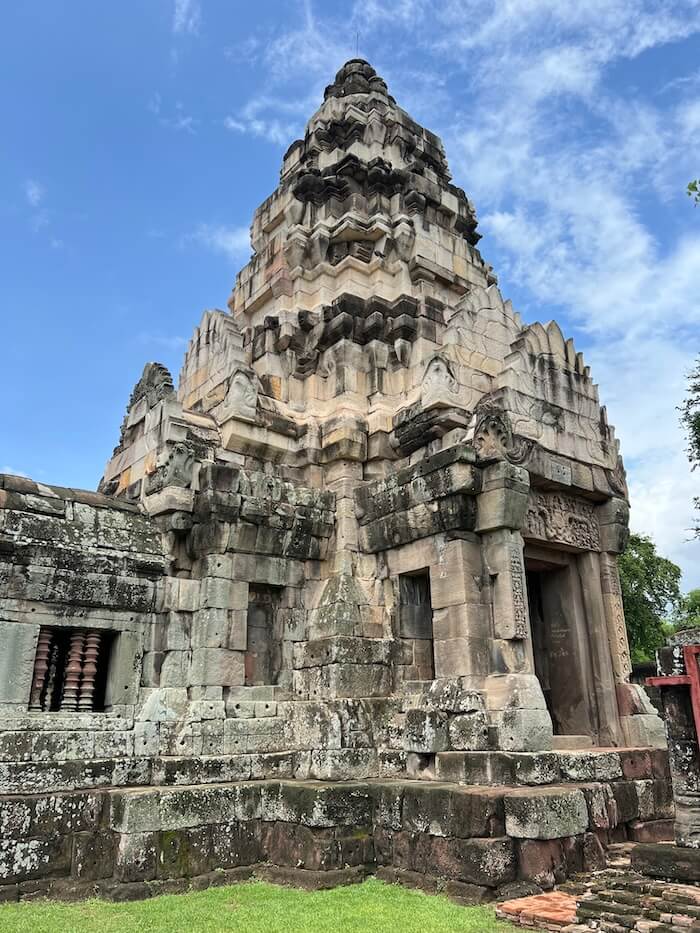 A tall, tiered sandstone and laterite Khmer sanctuary tower (prang) under a blue sky with light clouds, featuring intricate carvings above the doorway and traditional stone window balustrades.