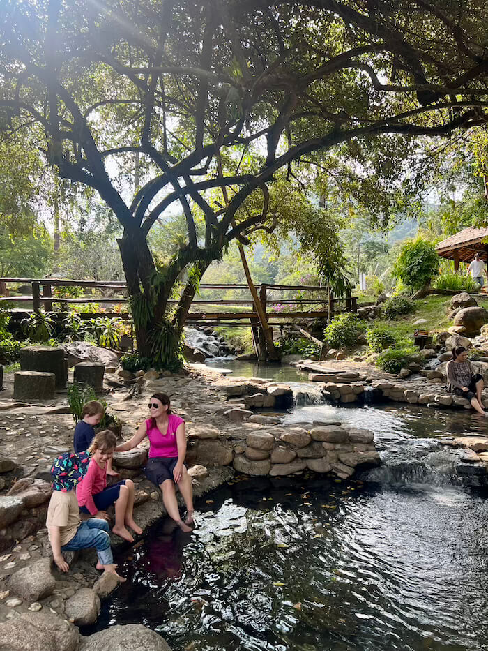 A woman and three young children sitting on large stones, soaking their feet in a clear mineral pool at Chae Son National Park.