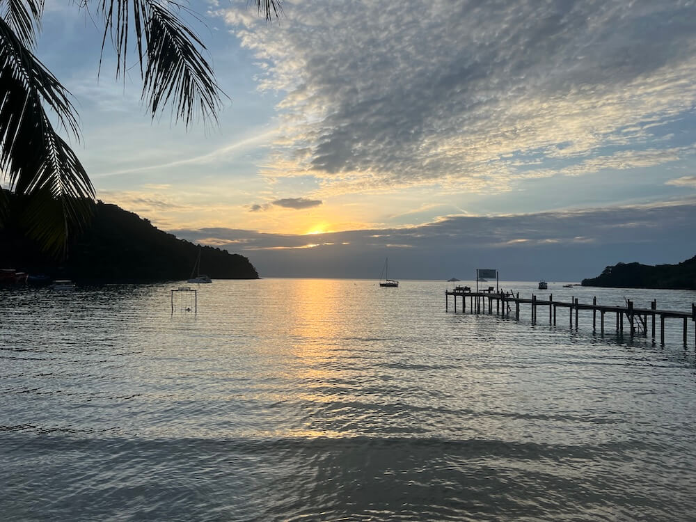 A peaceful sunset over a tropical bay in Koh Kood, Thailand, featuring a long wooden pier and sailboats.