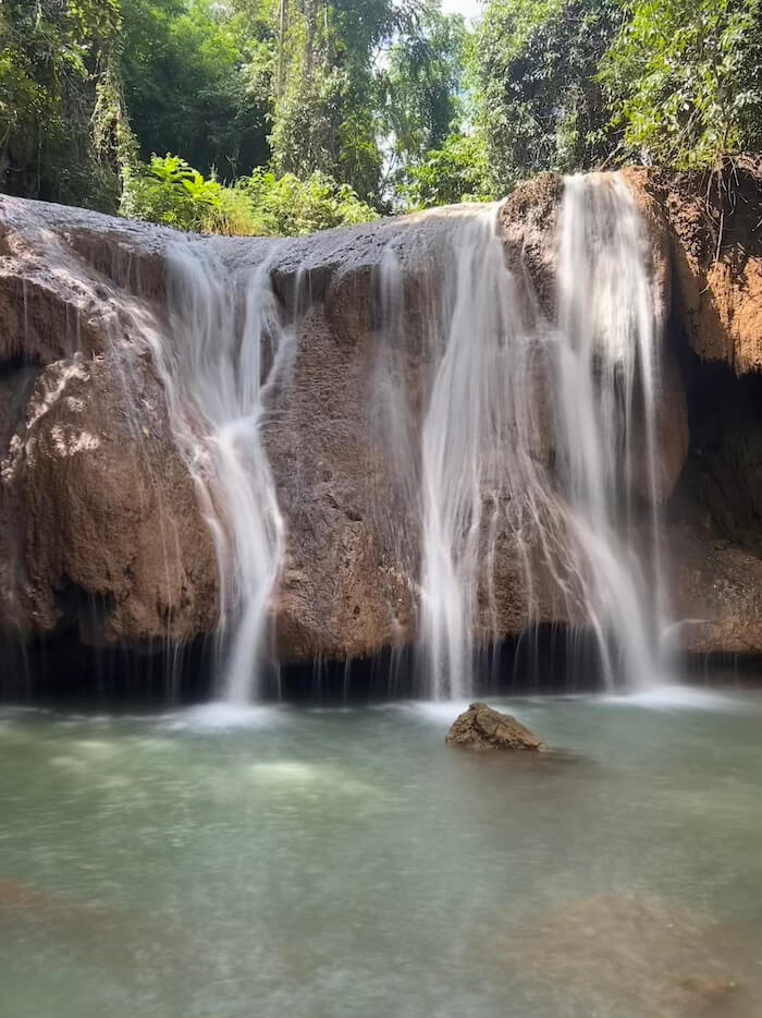 A close-up view of a wide, tiered waterfall in Doi Phu Nang National Park, with water cascading over smooth brown rock into a clear, sunlit turquoise pool.