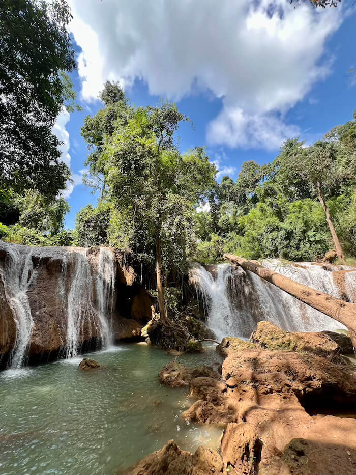 A wide-angle view of the multi-tiered Thansawan Waterfall under a bright blue sky with white clouds, showing various cascades and the surrounding dense green jungle.