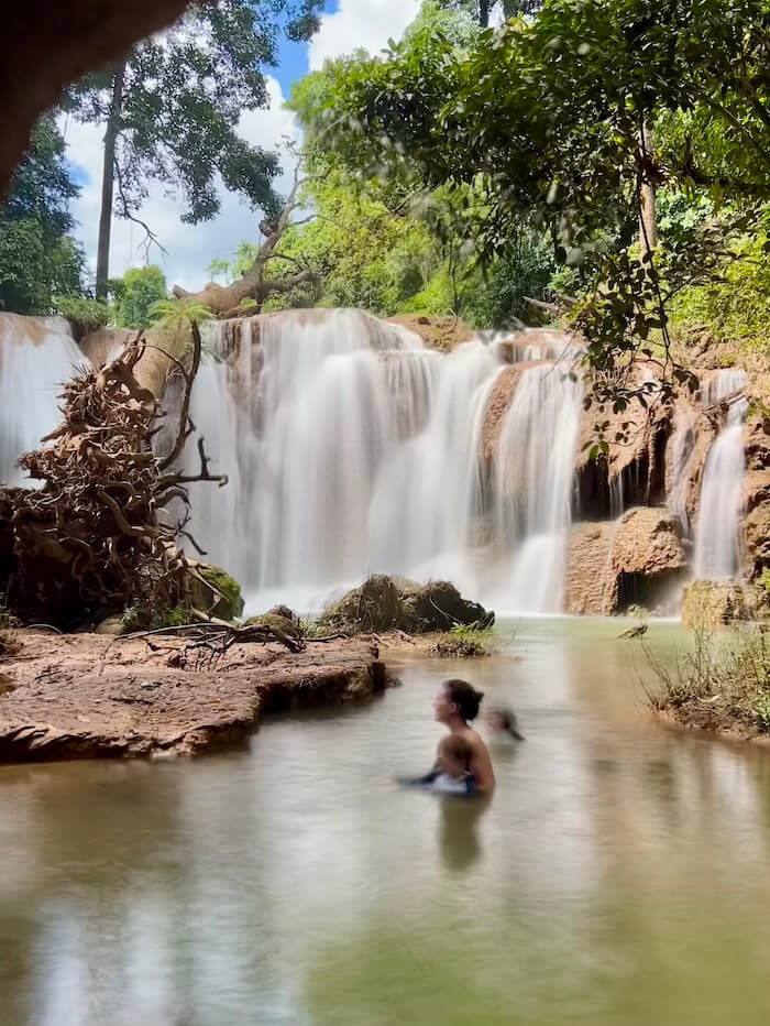A wide, silky long-exposure shot of Thansawan Waterfall in Thailand, featuring turquoise pools, lush green jungle, and a large fallen tree with dramatic roots in the foreground.