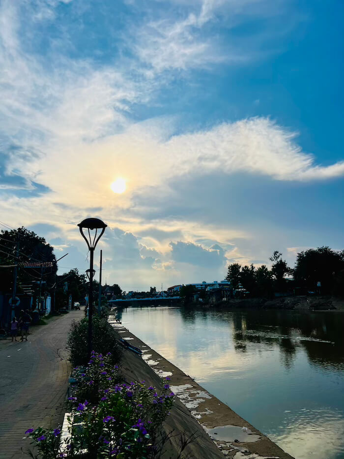 Wang River Riverside Path at Sunset A wide view of the Wang River in Lampang at sunset, with a brick walking path, street lamps, and purple flowers in the foreground and a soft sun glowing through clouds.