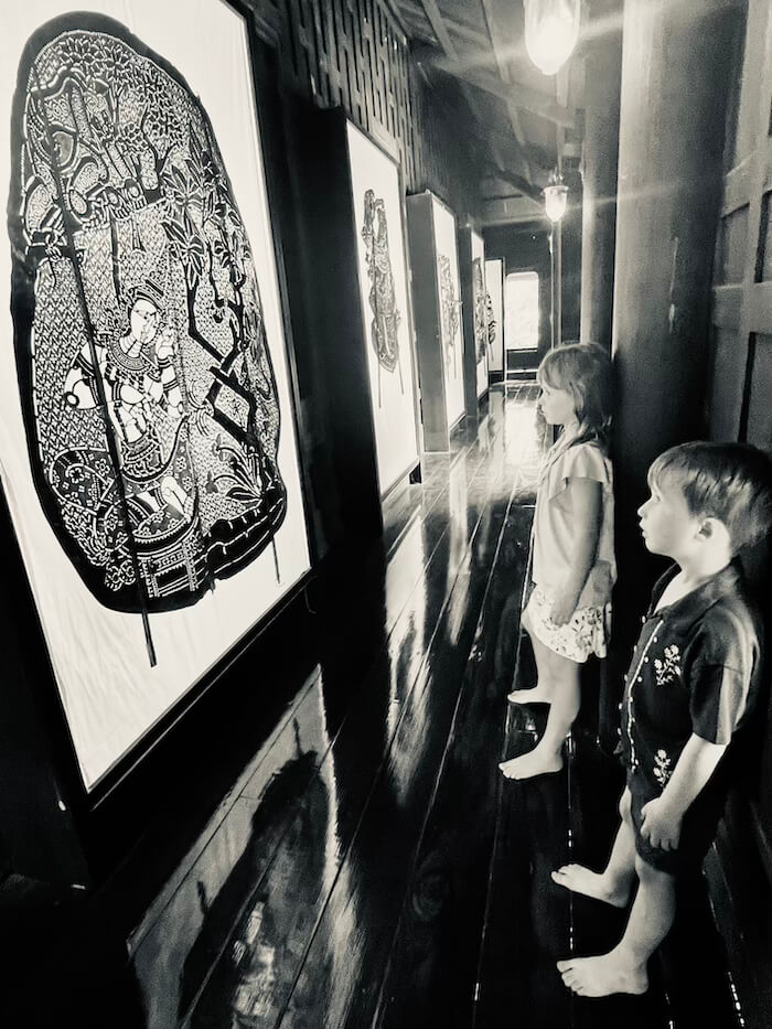 Black and white photo of two children looking at large, intricate Nang Yai shadow puppets displayed in a dimly lit museum with polished wooden floors.