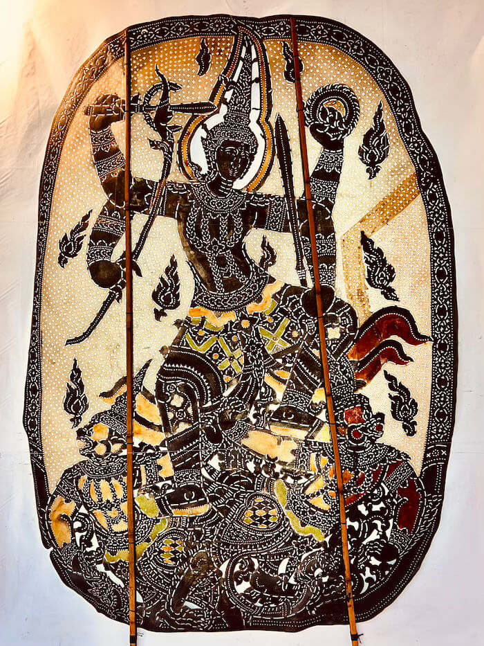 A close-up shot of a large, intricate Thai shadow puppet carved from cowhide, featuring a central figure from the Ramayana with multiple arms and weapons.