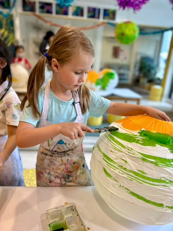 A young girl in a blue shirt and white apron carefully painting a large paper lantern with green paint.