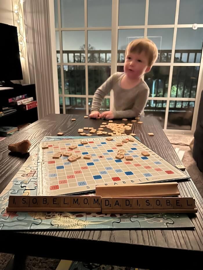 A young boy standing behind a table with a Scrabble board and letter tiles spelling out family names.