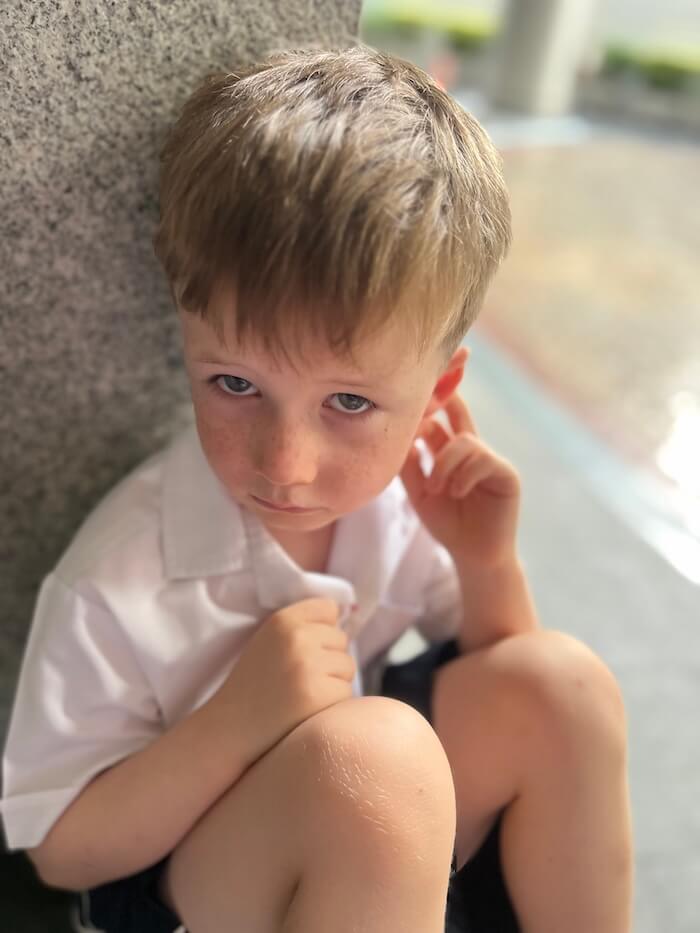A young boy with freckles and blond hair, wearing a white collared shirt, sitting pensively and looking directly at the camera with a thoughtful expression.