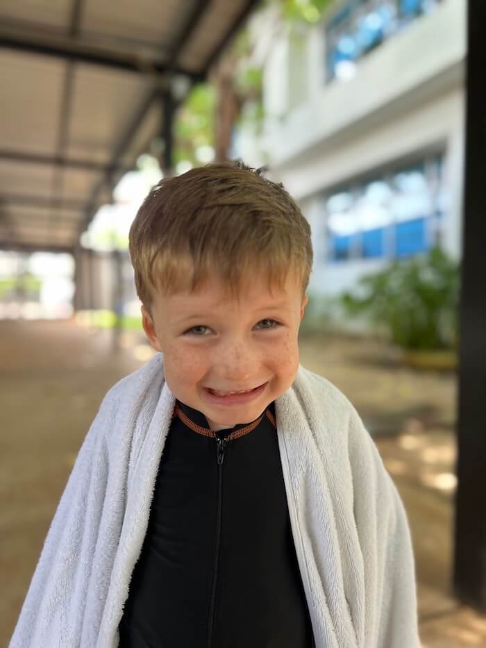 A young boy named Henry with freckles and a wide, cheeky grin, wrapped in a white towel over a black swim shirt.