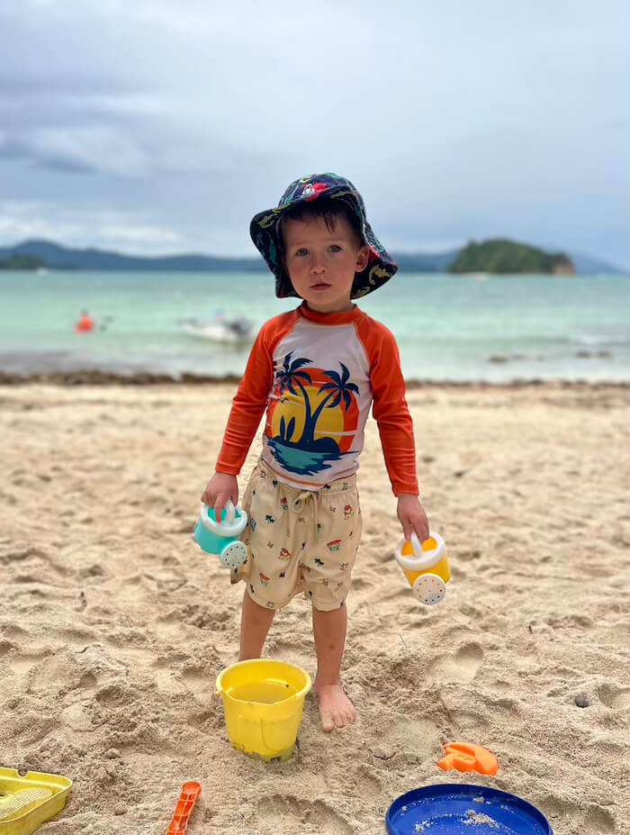 A young boy named Charlie standing on a sandy beach wearing an orange rash guard and a sun hat, holding two small watering cans with a yellow bucket at his feet.