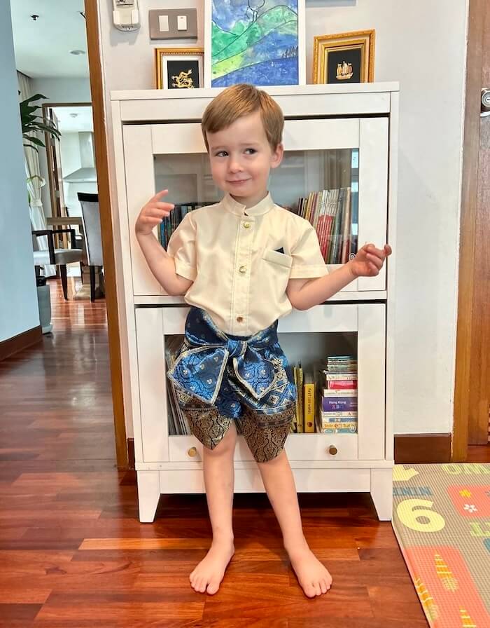 A young boy standing proudly in a white traditional Thai shirt and ornate blue and gold patterned wrap, smiling in an indoor setting.