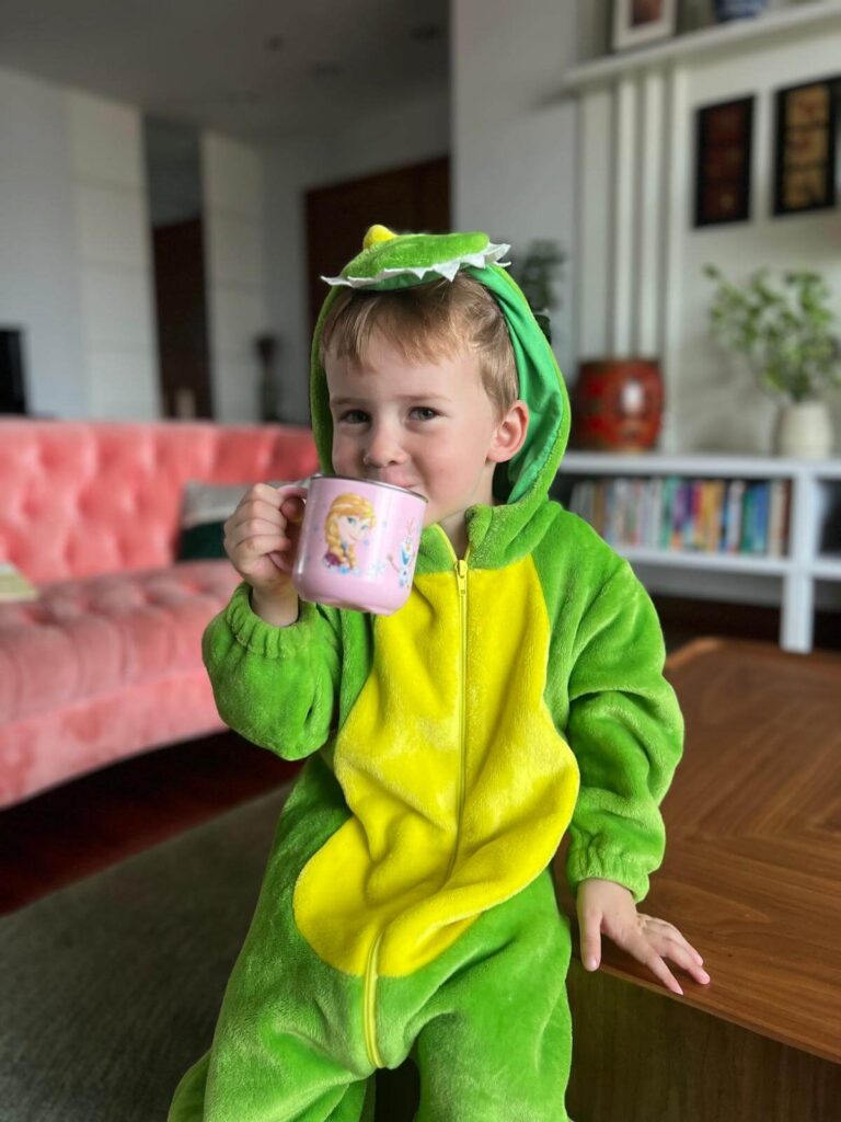 A young boy wearing a bright green dinosaur onesie, sitting on a wooden table and sipping from a pink mug with cartoon characters.