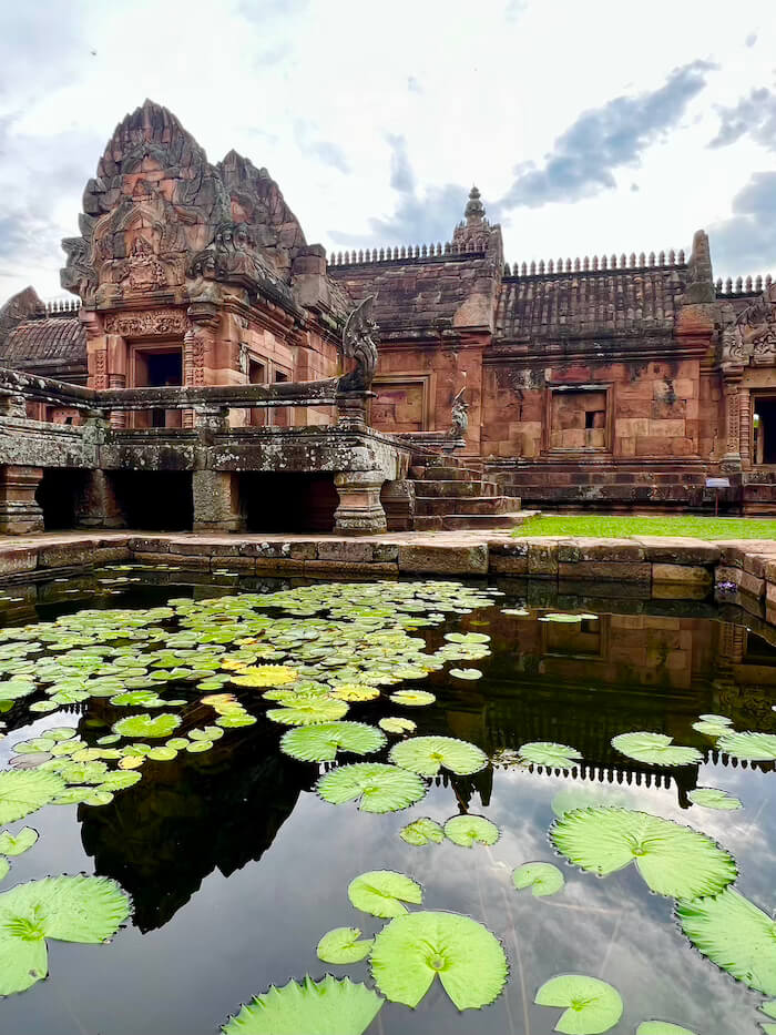 A wide shot of an ancient red sandstone temple with intricate carvings, reflected in a still pond covered in green lily pads.