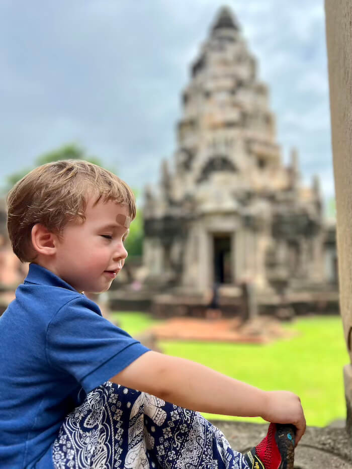 A young boy in a blue shirt and patterned pants sitting in front of the ancient stone prang of Phimai Historical Park.