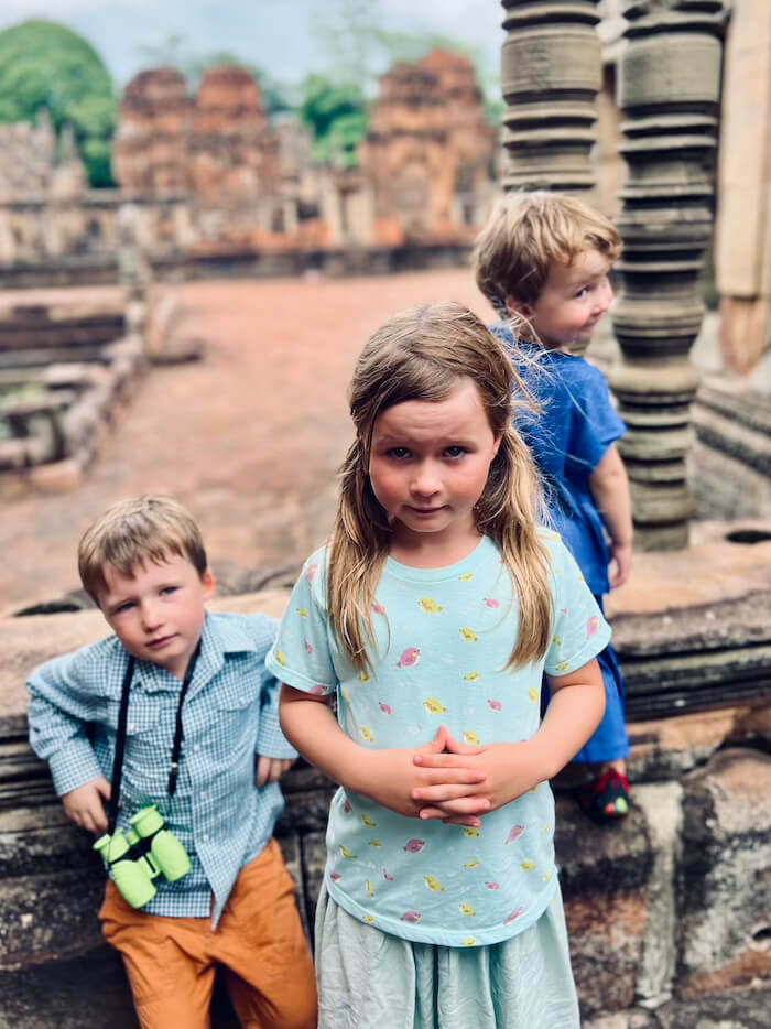 Three young children posing in front of ancient sandstone temple ruins with a mix of curious and tired expressions.