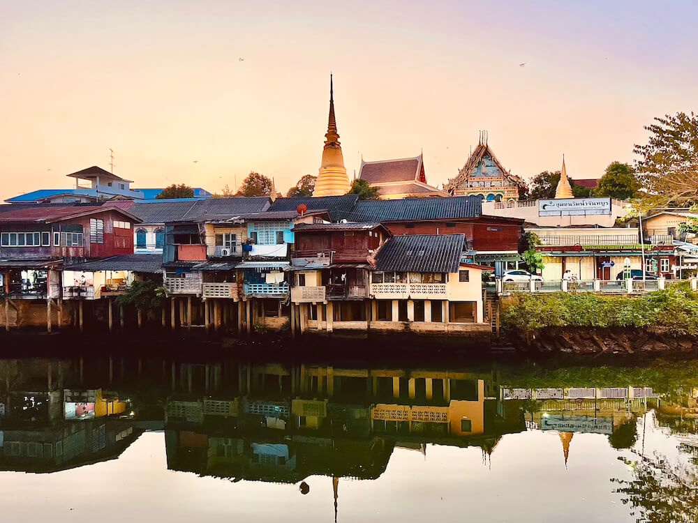 Historic wooden shophouses along the Chanthaburi River at sunset with the spires of the Cathedral of the Immaculate Conception in the background.