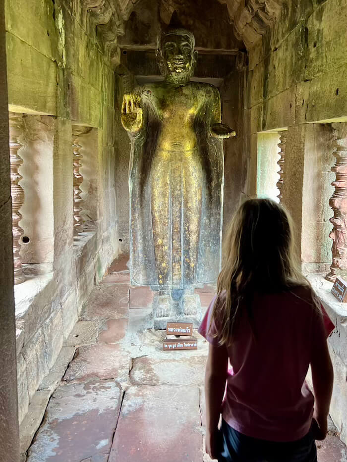 A young girl with long blonde hair seen from behind, looking at a large, ancient standing Buddha statue inside a stone corridor at Phimai Historical Park.