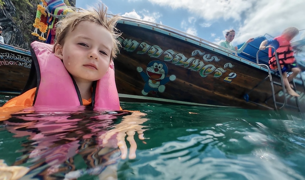 A boy in a pink lifejacket floats beside a longtail boat, about to begin a swim through the Emerald Cave.