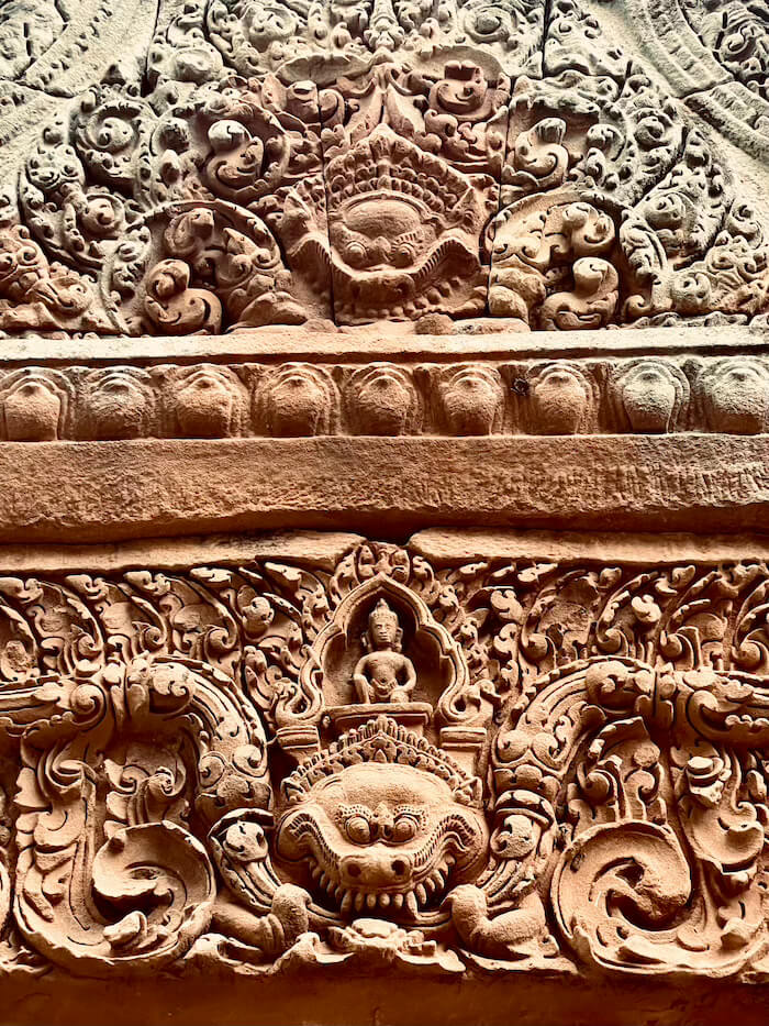 A close-up of an intricate stone carving on a temple lintel at Prasat Muang Tam, featuring the Kirtimukha (Kala Mask) with a fearsome face and bulging eyes, topped by a small seated deity.