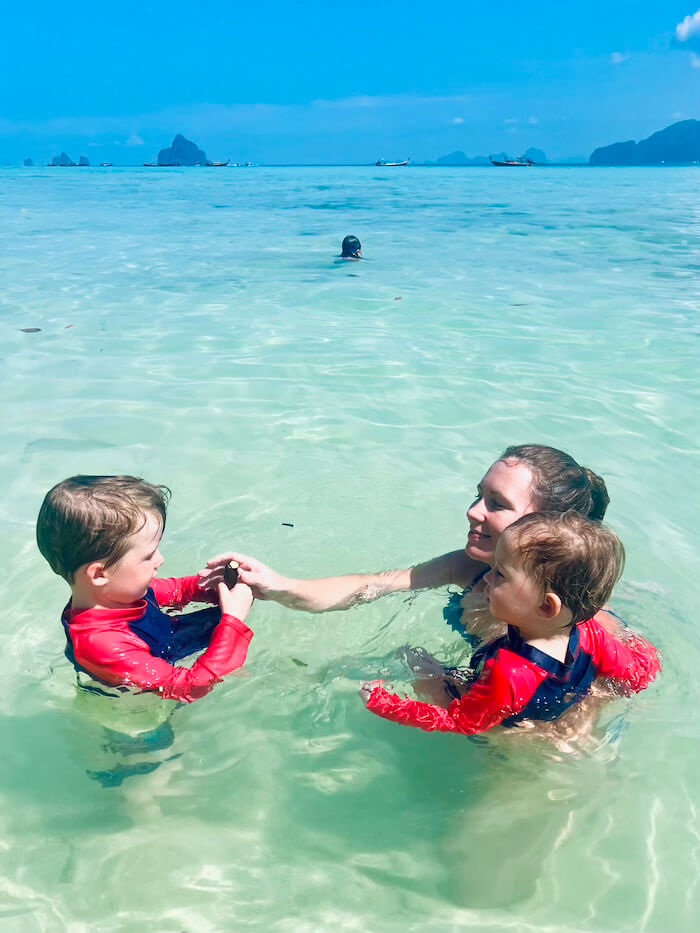 A mother and two young children in red sunshirts playing in waist-deep, crystal-clear turquoise water at Koh Kradan, with a bright blue sky and distant islands.