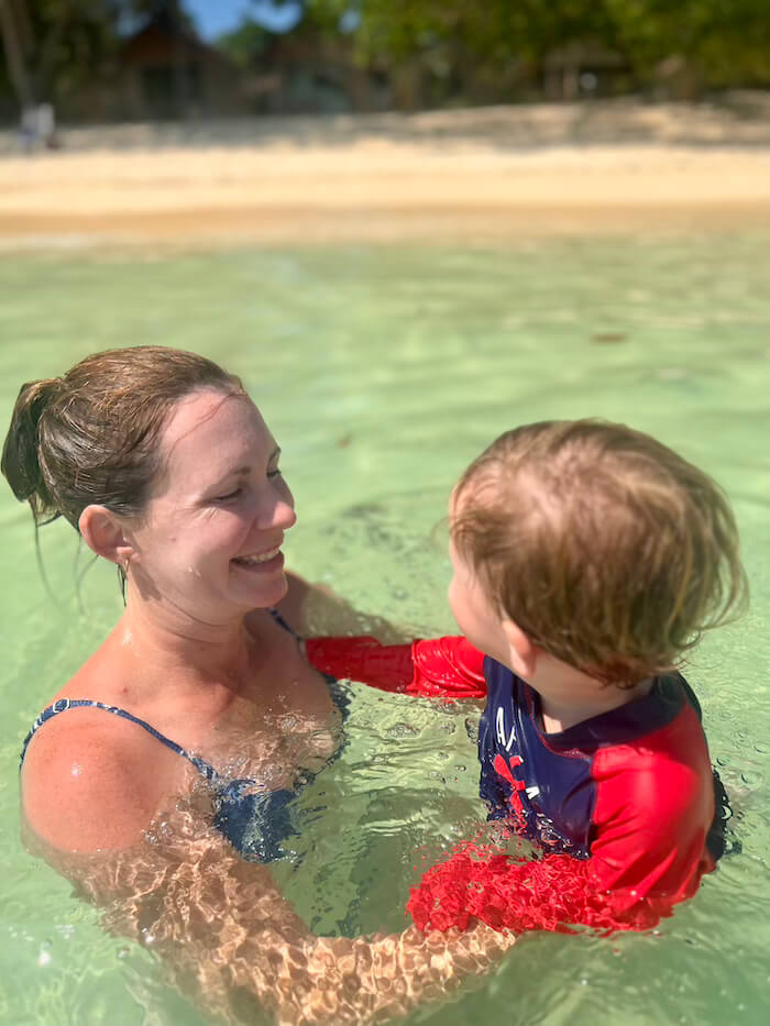 A mother and young son smiling at each other while swimming in chest-deep, crystal-clear turquoise water at a tropical beach.