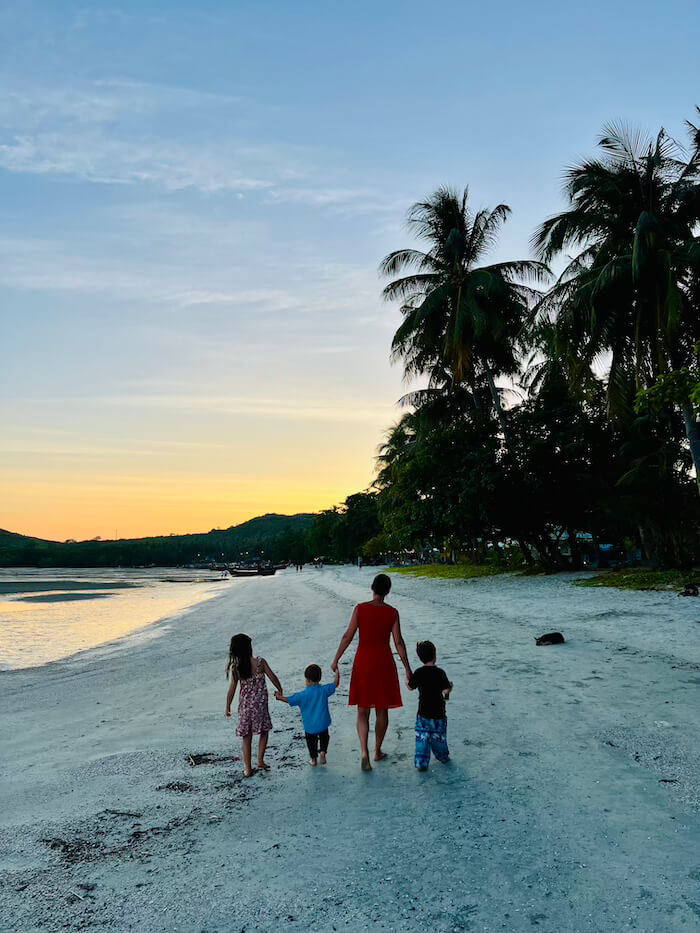 Silhouette of a mother and three children walking hand-in-hand along a white sand beach at sunset, framed by tall coconut palms on Koh Muk.