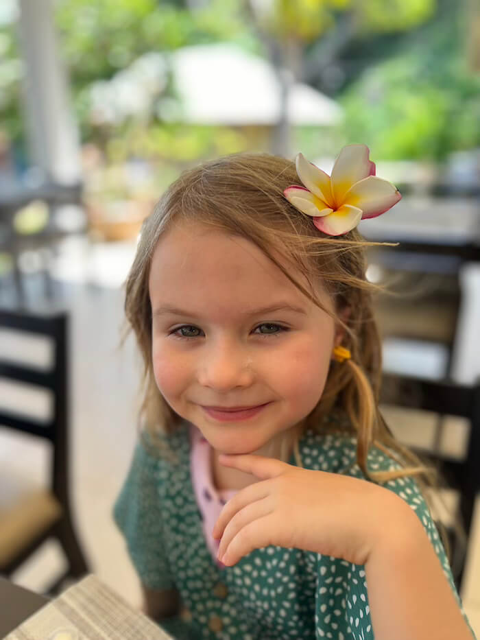 Close-up portrait of a smiling young girl with a pink and white frangipani flower in her hair at an outdoor restaurant.