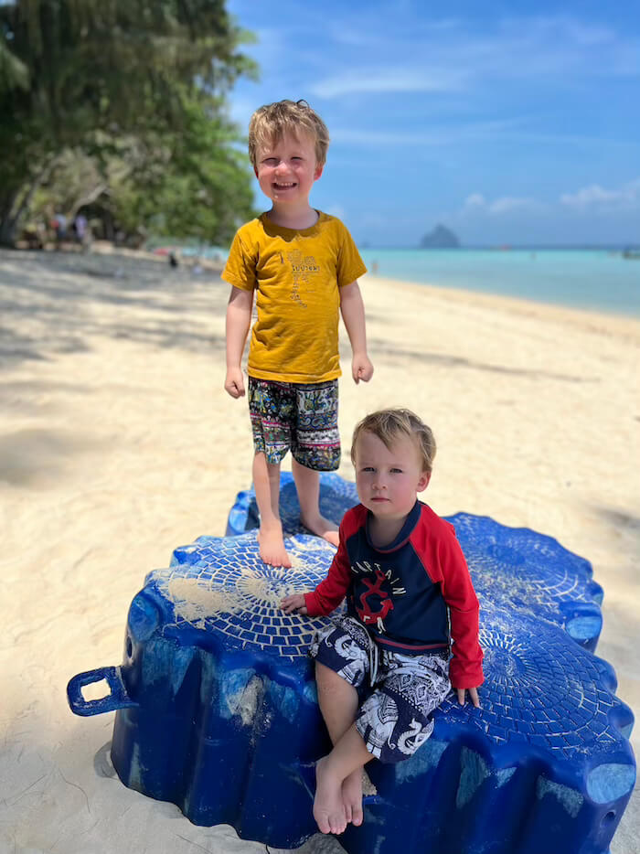 Two young boys, one in a yellow t-shirt and the other in a red and blue sunshirt, smiling while standing on a blue plastic dock on a white sand beach at Koh Ngai.