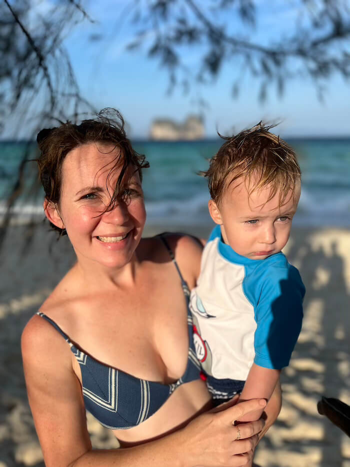 A mother smiling and holding her young son on a beach at Koh Ngai during golden hour, with the ocean and a limestone island in the blurred background.