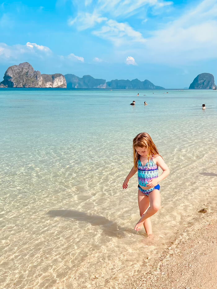 A young girl in a blue swimsuit walking through shallow, transparent turquoise water on Koh Ngai beach with a large limestone island in the background.