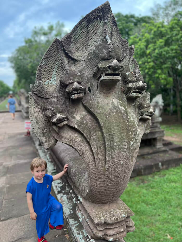A young boy in blue standing next to a massive, five-headed stone Naga serpent sculpture at the start of the naga bridge at Phanom Rung Historical Park.