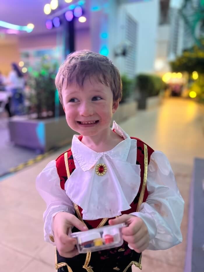 A smiling young boy dressed in an ornate red and white costume with a gold brooch, holding a small plastic container.