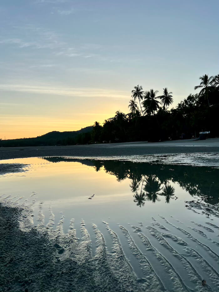 A vertical shot of a golden sunset reflected in a calm tidal pool on a rippled sandy beach at Koh Muk, Thailand, with silhouettes of palm trees.