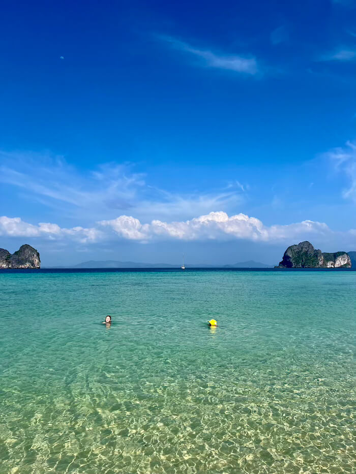 Two children swimming in vibrant, transparent turquoise water at Koh Ngai with dramatic limestone karsts on the horizon under a deep blue sky.
