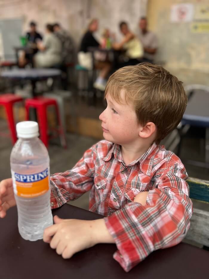 A young boy in a red plaid shirt sitting at an outdoor table, looking off to the side with a curious and engaged expression.